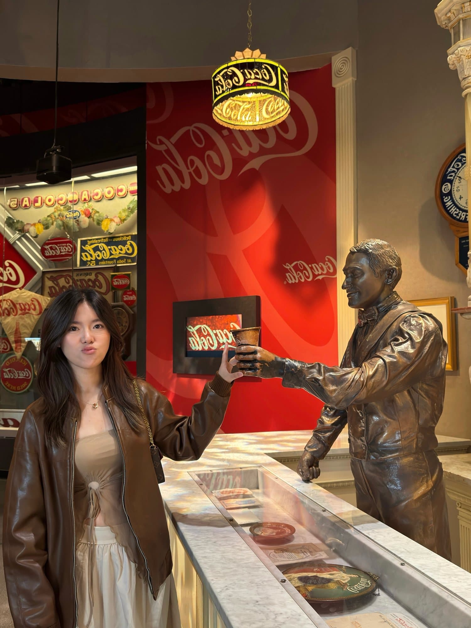 In Atlanta at the Coca-Cola museum, posing with a bronze statue that holds out a cup in front of red Coca-Cola signs