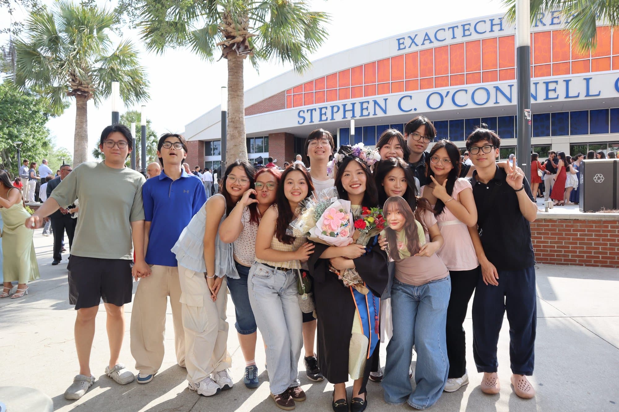 A big group of friends gathered around a graduate holding bouquets, in front of the Stephen C. O’Connell Center