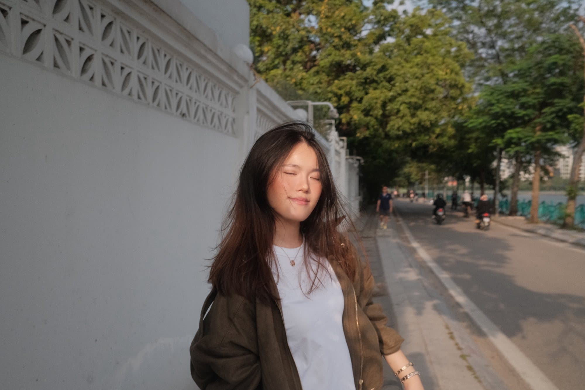 A woman with her eyes closed in warm evening light beside a white lattice wall on a tree-lined Hanoi lakeside road with motorbikes