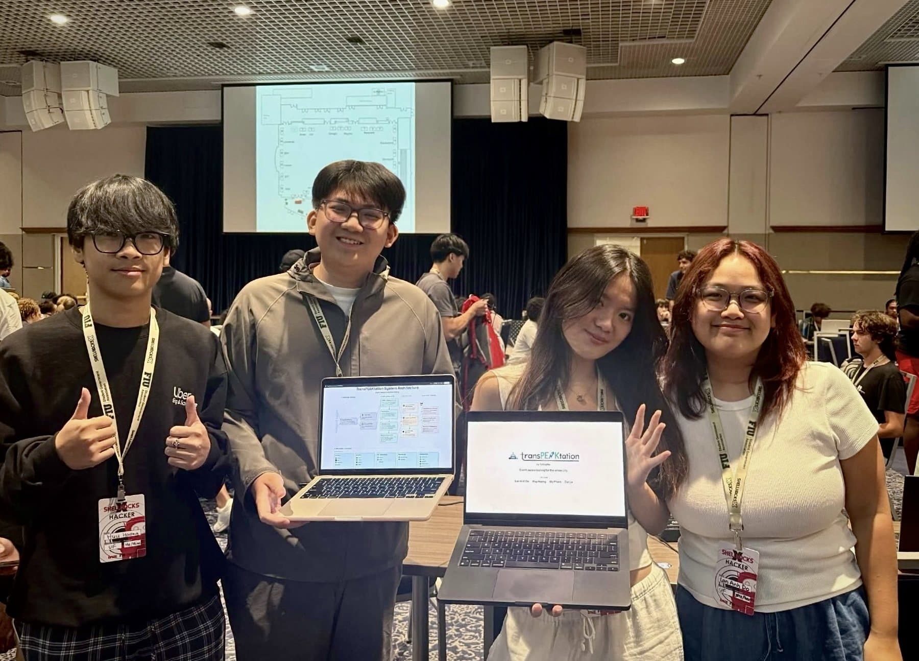The transPEAKtation team at ShellHacks holding laptops that show the project's architecture diagram and title screen
